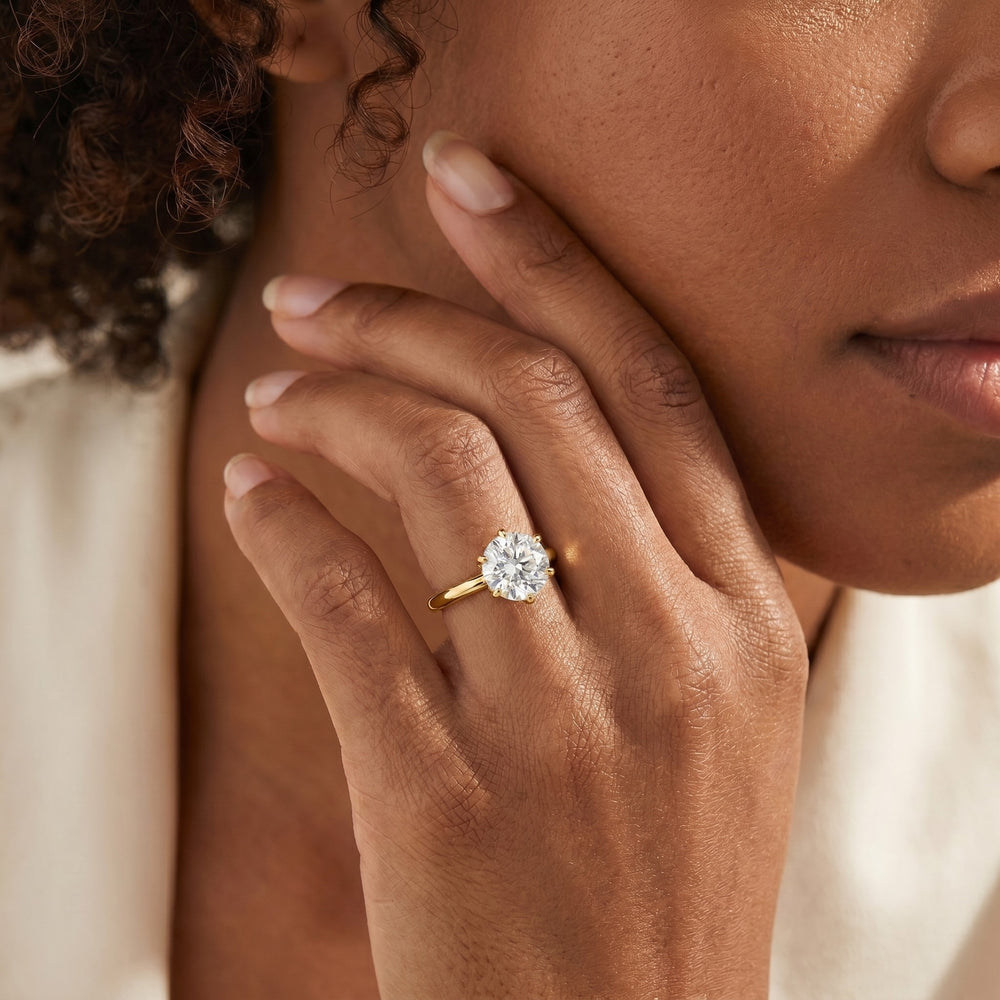 Close-up of a person touching their face, showcasing a gold ring with a large, sparkling diamond. The image conveys elegance and luxury.