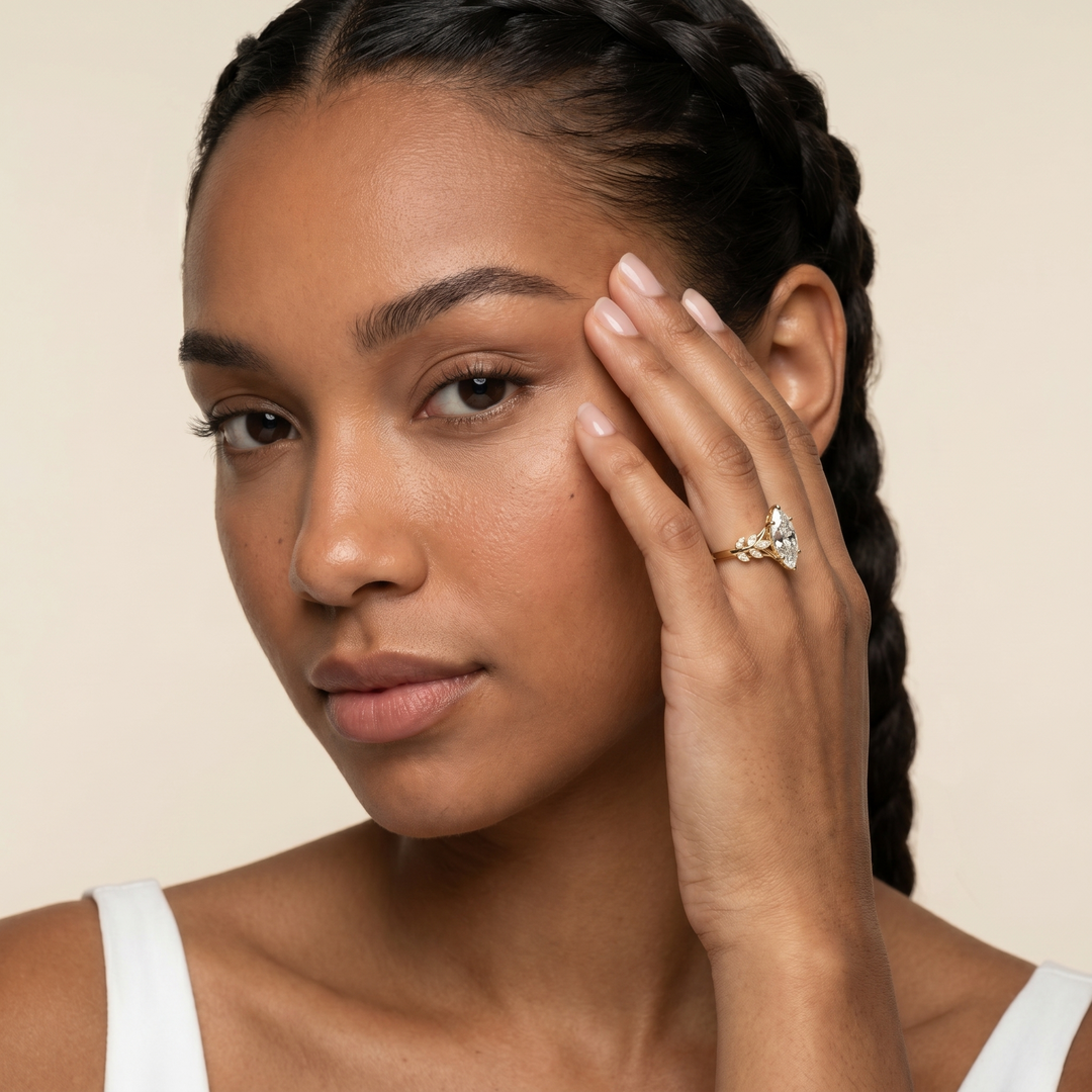A woman with braided hair gently touches her face, showcasing a gold ring with a large gem, conveying elegance and sophistication against a neutral background.