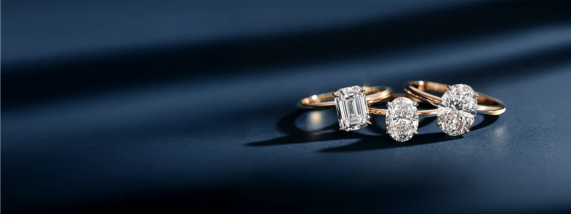 An elegant banner image showcasing a collection of gold solitaire engagement rings with different fancy-shaped diamonds, including emerald and oval cuts, on a navy blue background.