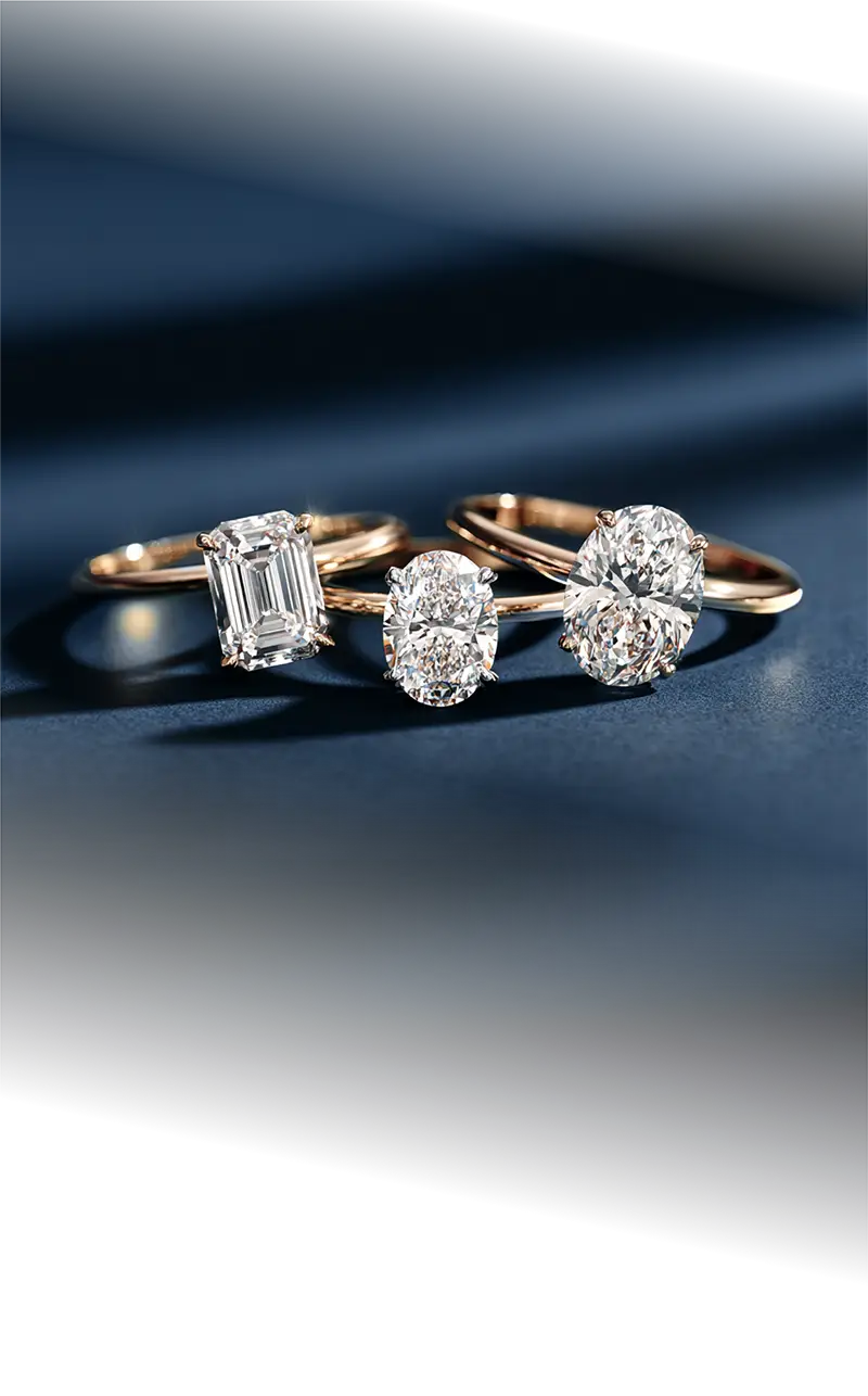 A vertical photograph showcasing a collection of three gold solitaire engagement rings with different fancy diamond shapes, including an emerald cut and two oval cuts, on a navy blue background.