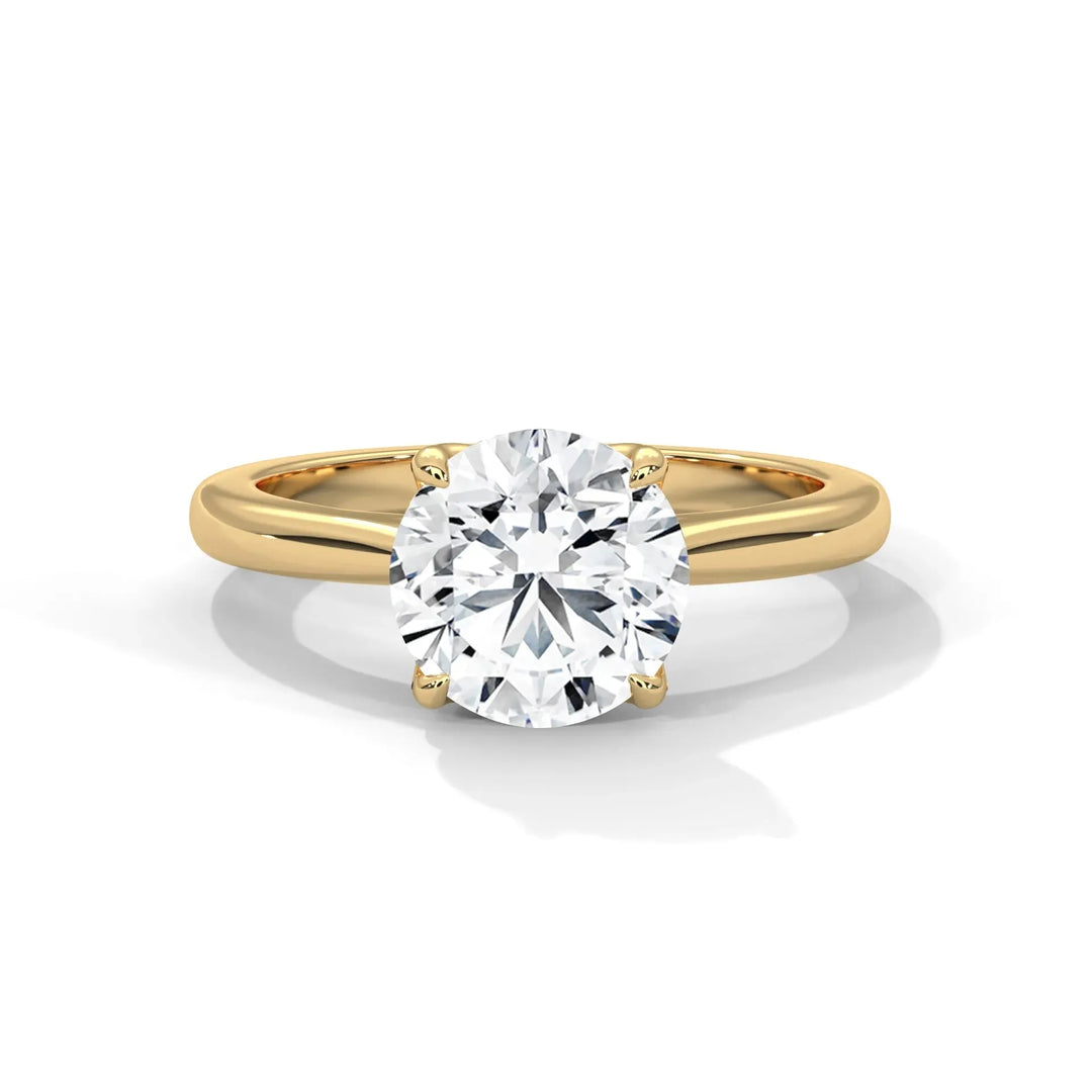 A gold band solitaire ring features a large, round diamond secured by four prongs. The reflection on a white surface evokes elegance and sophistication.