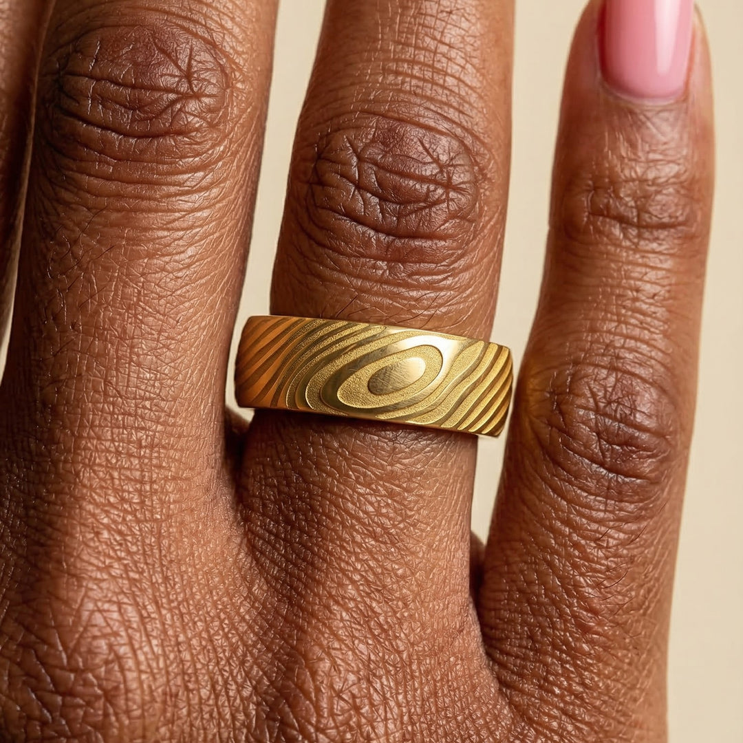 Gold ring with wood grain pattern on a person's finger against a beige background