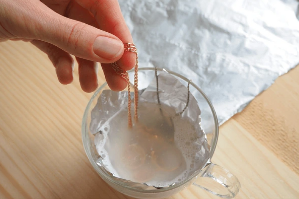 A hand holds a chain above a glass bowl containing a silver cleaning solution, with aluminum foil inside the bowl.