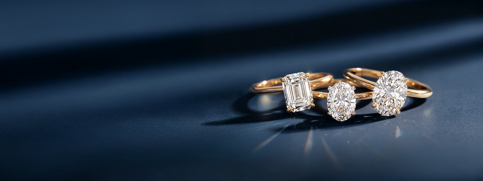 Three gold diamond rings rest on a dark blue background, elegantly illuminated. The rings feature emerald, oval, and round brilliant-cut diamonds.