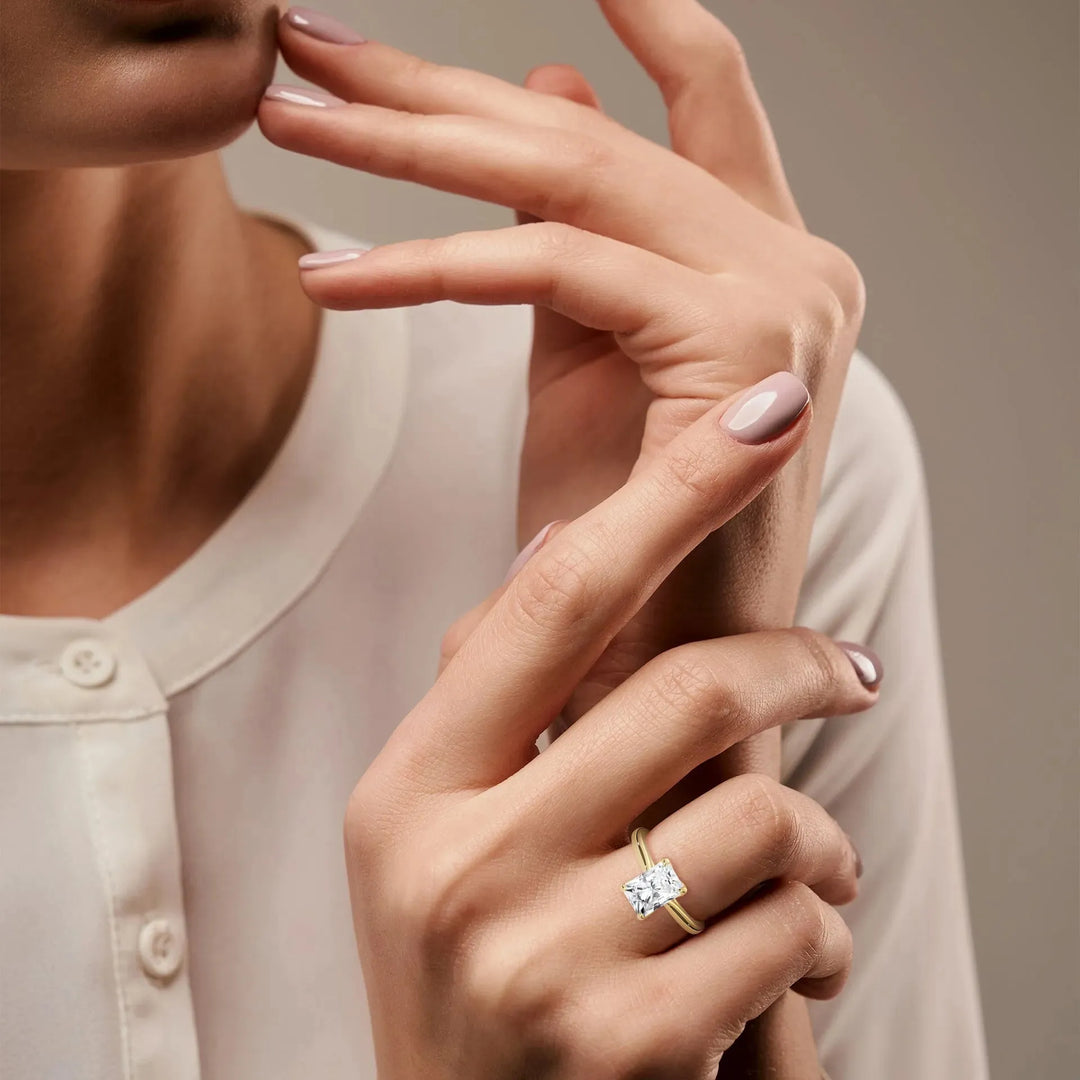 Woman's hands with light pink nail polish, one wearing a gold ring with a large square diamond. She is wearing a white blouse, displaying elegance.